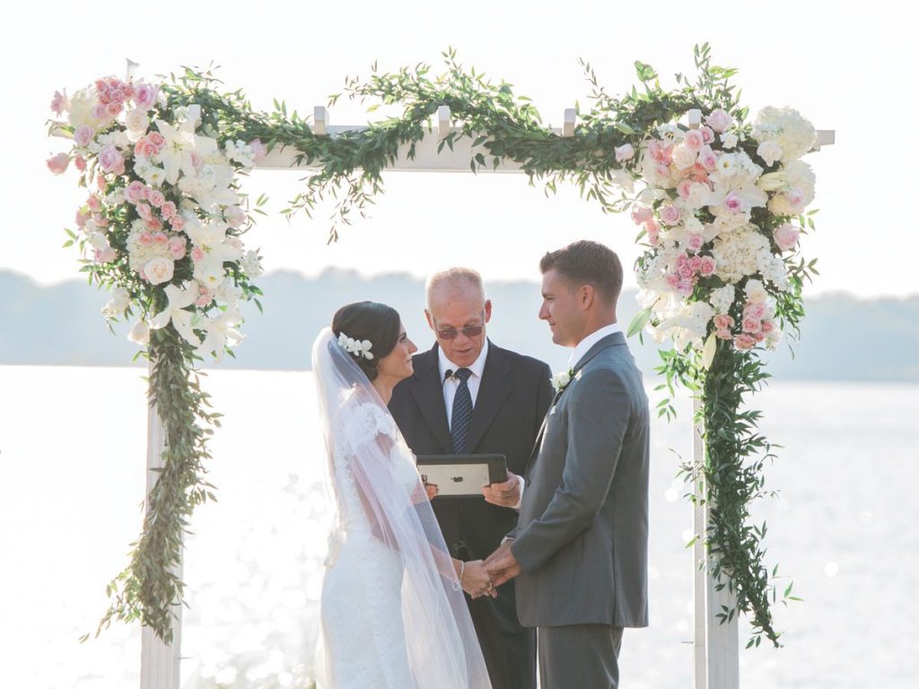Wedding ceremony taking place at a beach. Bride and groom facing each other holding hands in front of a minister as he recites a wedding ceremony script.