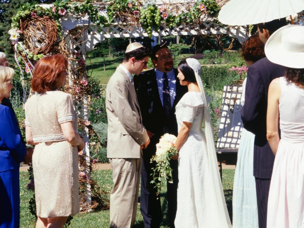 Jewish wedding ceremony taking place in an outdoor setting. With friends and family by the sides of the bride and groom under a flower decorated altar.
