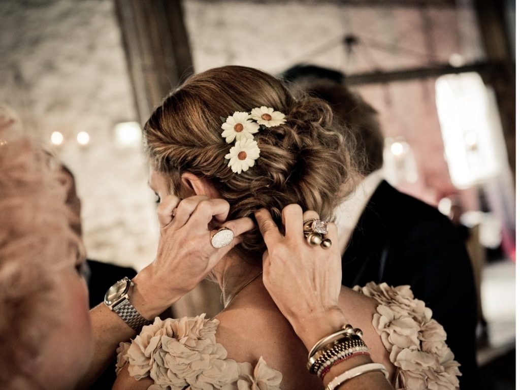 Woman facing away while another woman fixes her hair. Hair is styled in rollup curls with flower accents.