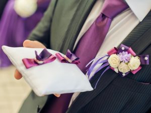 Close up of a ring bearer wearing a suit with a purple necktie holding a pillow with two rings tied to a purple ribbon