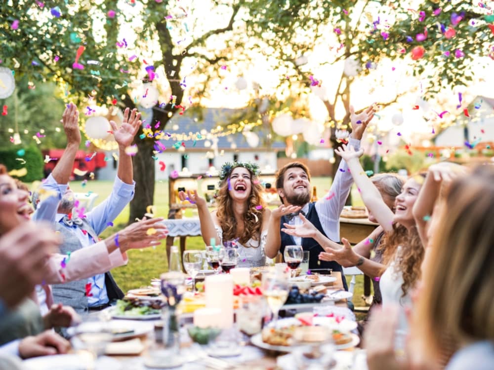 A couple dining outdoors with guests at the table and throwing confetti as wedding entertainment
