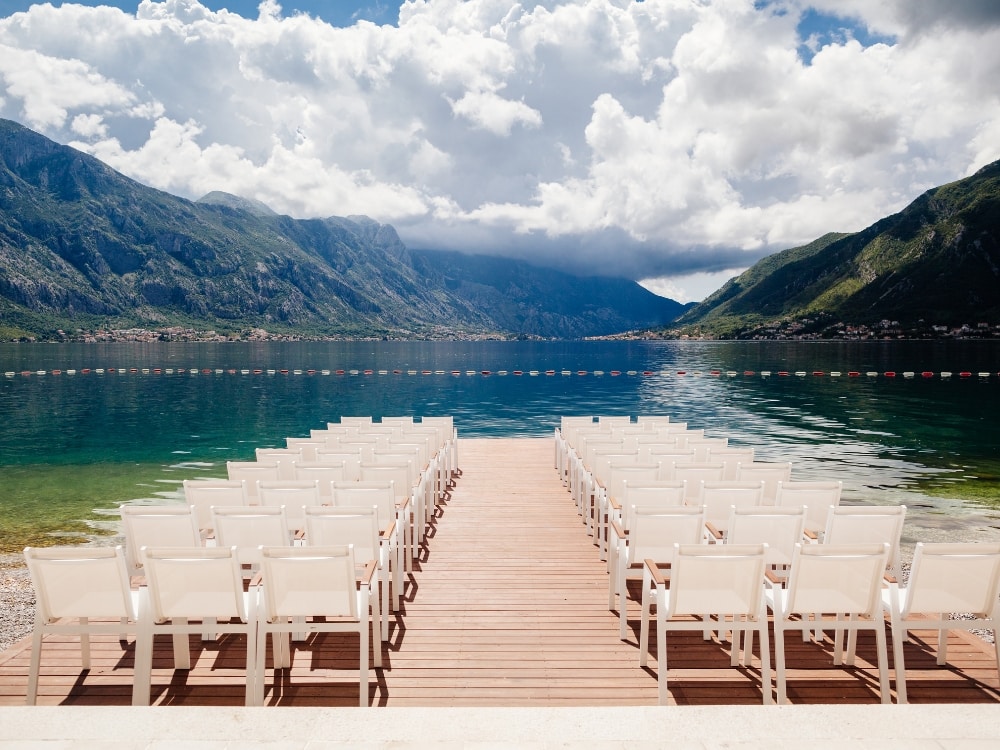 Chairs lined along a dock facing a lake slash mountain view.